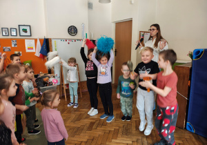 Trzy diewczynki stoją z pomponami uniesionymi ku górze. Dwóch chłopców stoi z instrumentami. Jeden chłopiec skacze. Za nimi stoi kobieta z mikrofonem. Przed nimi stoją przedszkolaki.