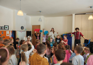 Trzy diewczynki stoją z pomponami uniesionymi ku górze. Dwóch chłopców stoi z instrumentami. Jeden chłopiec skacze. Za nimi stoi kobieta oraz mężczyzna z mikrofonem. Przed nimi stoją przedszkolaki.