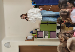 Kobieta w okularach stoi trzymając mikrofon. Przed nią siedzą przedszkolaki.