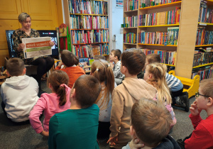 Przedszkolaki siedzą na dywanie. Przed nimi kobieta z planszą "Smok Wawelski". Po bokach regały z książkami.