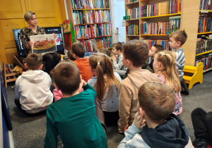 Przedszkolaki siedzą na dywanie. Przed nimi kobieta z planszą zamku. Po bokach regały z książkami.