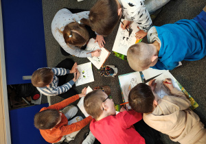 Przedszkolaki leżą na dywanie i kolorują smoki.