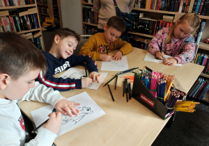 Przedszkolaki siedzą przy stole i kolorują smoki.