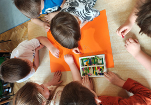 Przedszkolaki siedzą przy stole i malują pastelami na pomarańczowym kartonie.