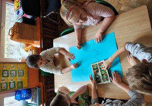 Przedszkolaki siedzą przy stole i malują pastelami na niebieskim kartonie.