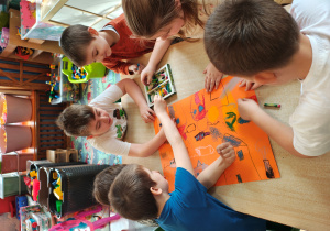 Przedszkolaki siedzą przy stole i malują pastelami na pomarańczowym kartonie.