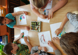 Przedszkolaki siedzą przy stole i kolorują smoka.