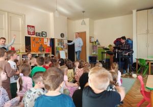 Przedszkolaki siedzą na dywanie. Przed nimi jest dwóch mężczyzn. Jeden stoi, drugi gra na perkusji elektrycznej.