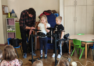 Dziewczynka i chłopiec stoją przy perkusji elektrycznej. Mężczyzna trzyma pałeczki do perkusji.