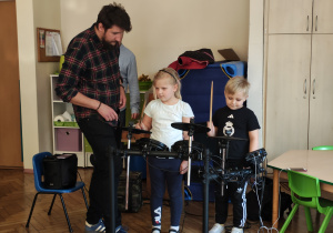 Dziewczynka i chłopiec stoją przy perkusji elektrycznej, trzymaja pałeczki do perkusji. Obok nich stoi mężczyzna.