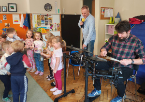 Pięć dziewczynek trzyma instrumenty. Za nimi stoi mężczyzna z mikrofonem. Obok mężczyzna gra na perkusji elektrycznej. Przed nimi skaczące dzieci.