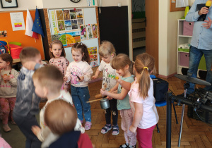 Pięć dziewczynek trzyma instrumenty. Za nimi stoi mężczyzna z mikrofonem. Obok stoi perkusja elektryczna. Przed nimi skaczące dzieci.