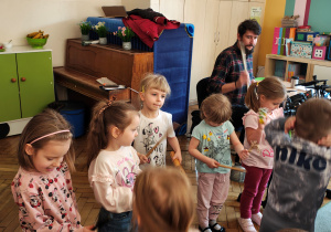 Pięć dziewczynek trzyma instrumenty. Obok mężczyzna gra na perkusji elektrycznej. Przed nimi skaczące dzieci.