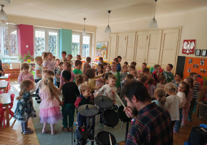 Pięć dziewczynek trzyma instrumenty. Obok mężczyzna gra na perkusji elektrycznej. Przed nimi skaczące dzieci.